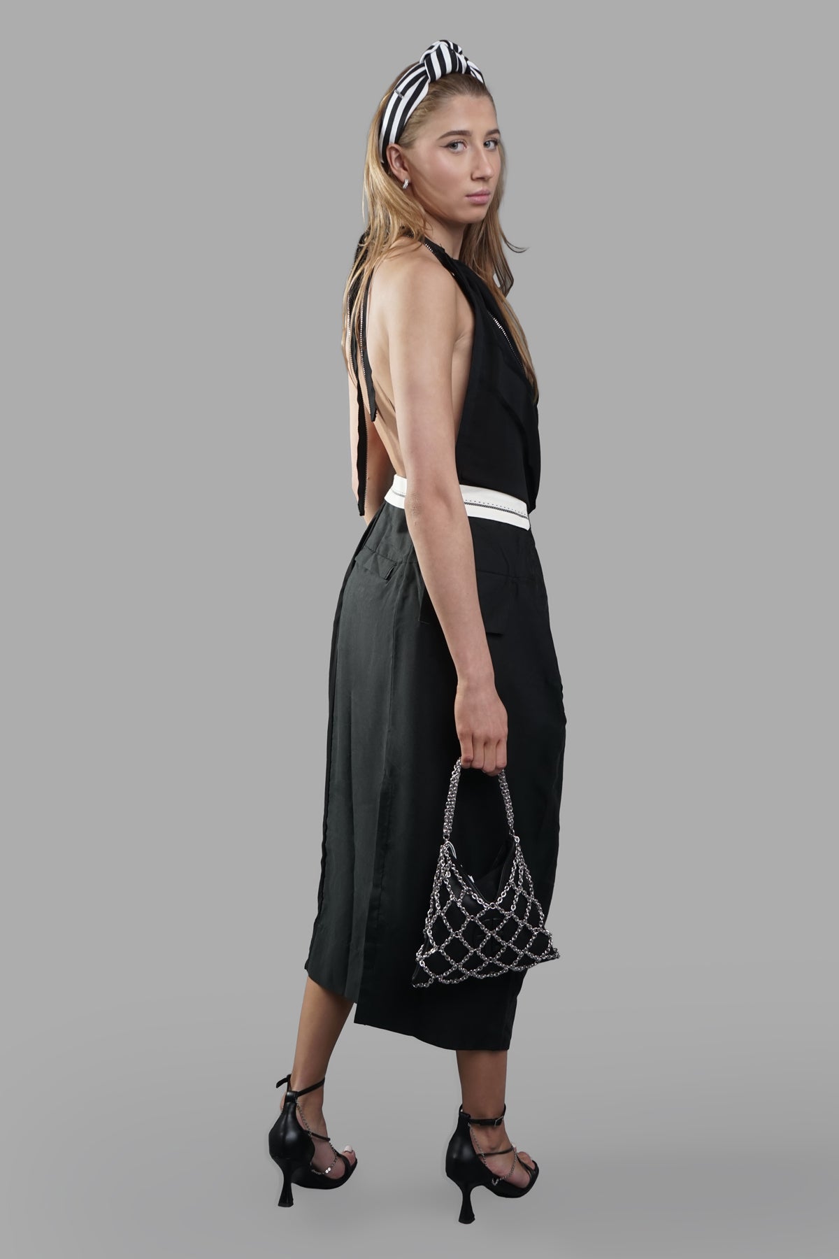Giovanni Cavagna open-back dress in black, featuring a halterneck and draped front. Side view on model with chic handbag.