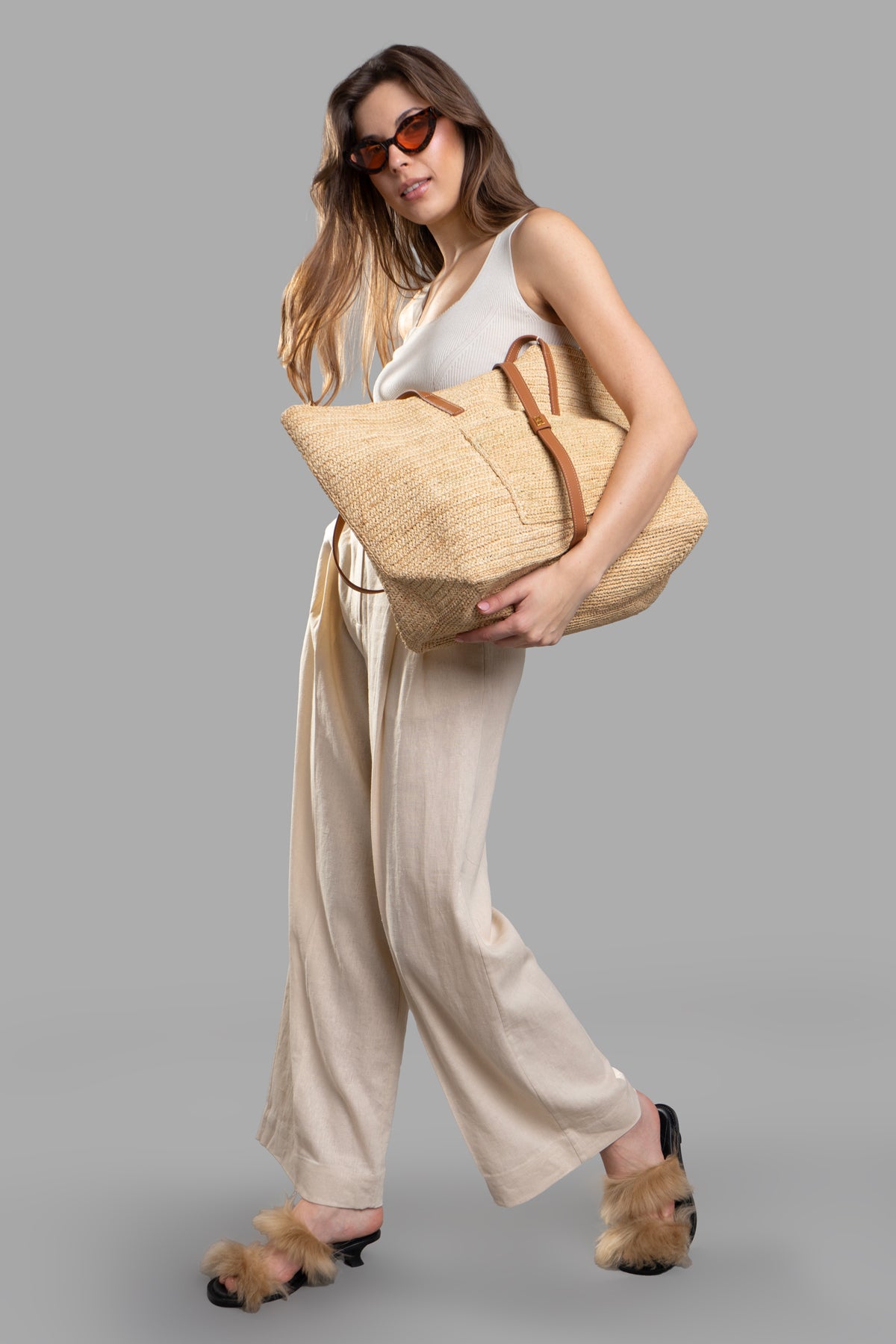 By Malene Birger high-waisted trousers, flared wide legs, beige, on model with sunglasses and woven tote bag.