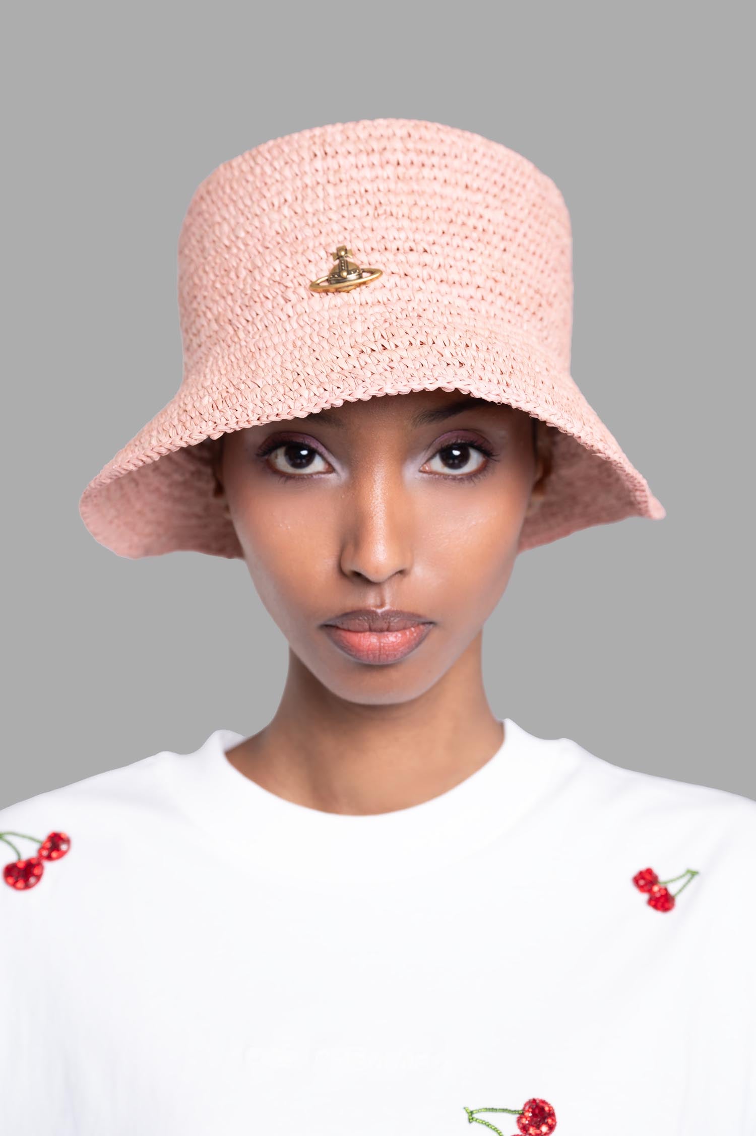 Vivienne Westwood Orb Raffia Bucket Hat in pink, featuring a wide brim and raised orb plaque, worn by model against a grey.