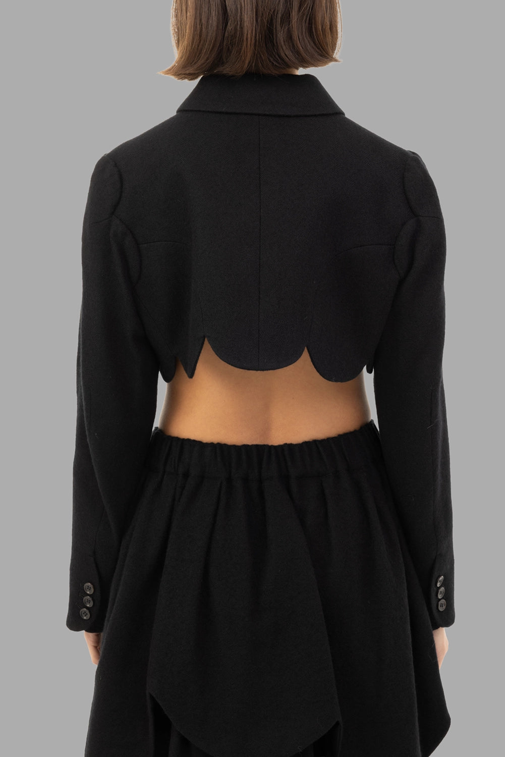 Noir Kei Ninomiya women’s cropped wool jacket in black, back view, open back with scalloped hem and button cuffs