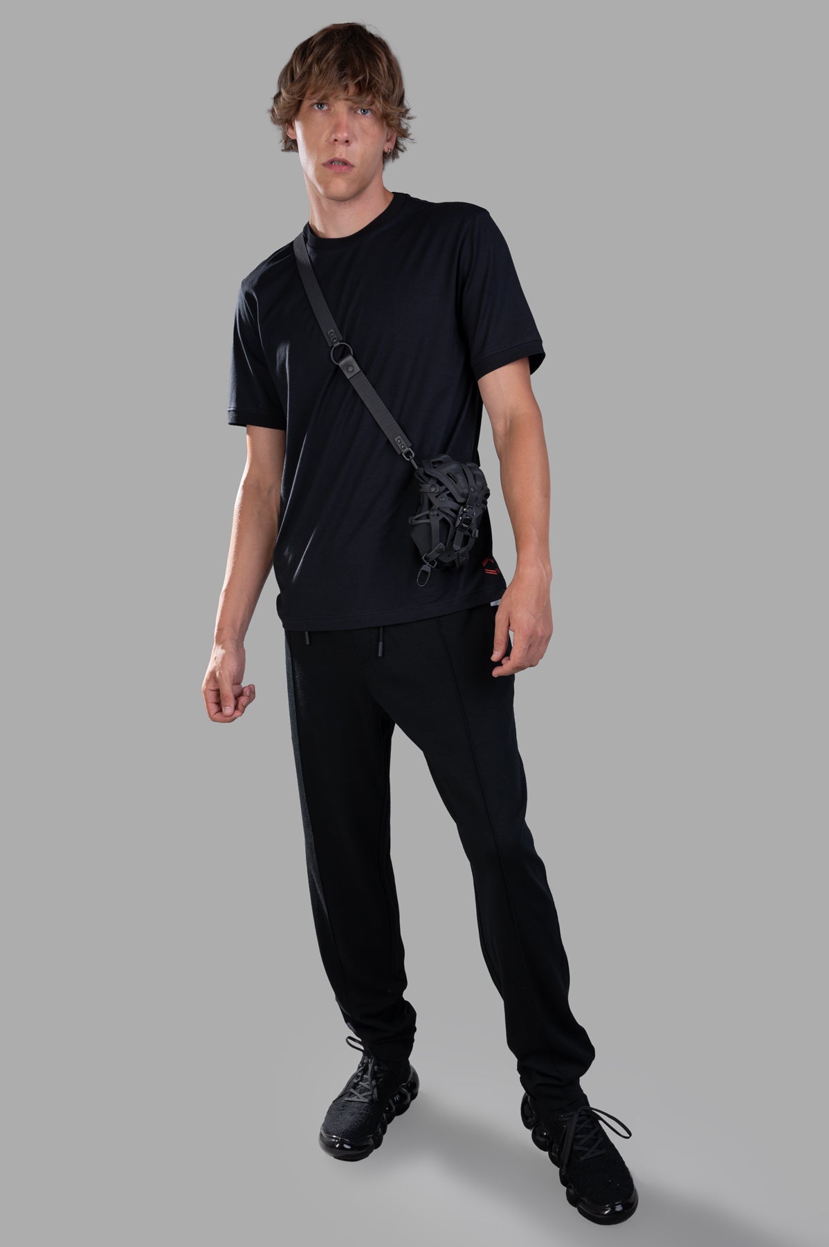 Orlebar Brown 'Lamborghini' Logo T-shirt on-model front view, black tee with black pants and crossbody bag, grey background.