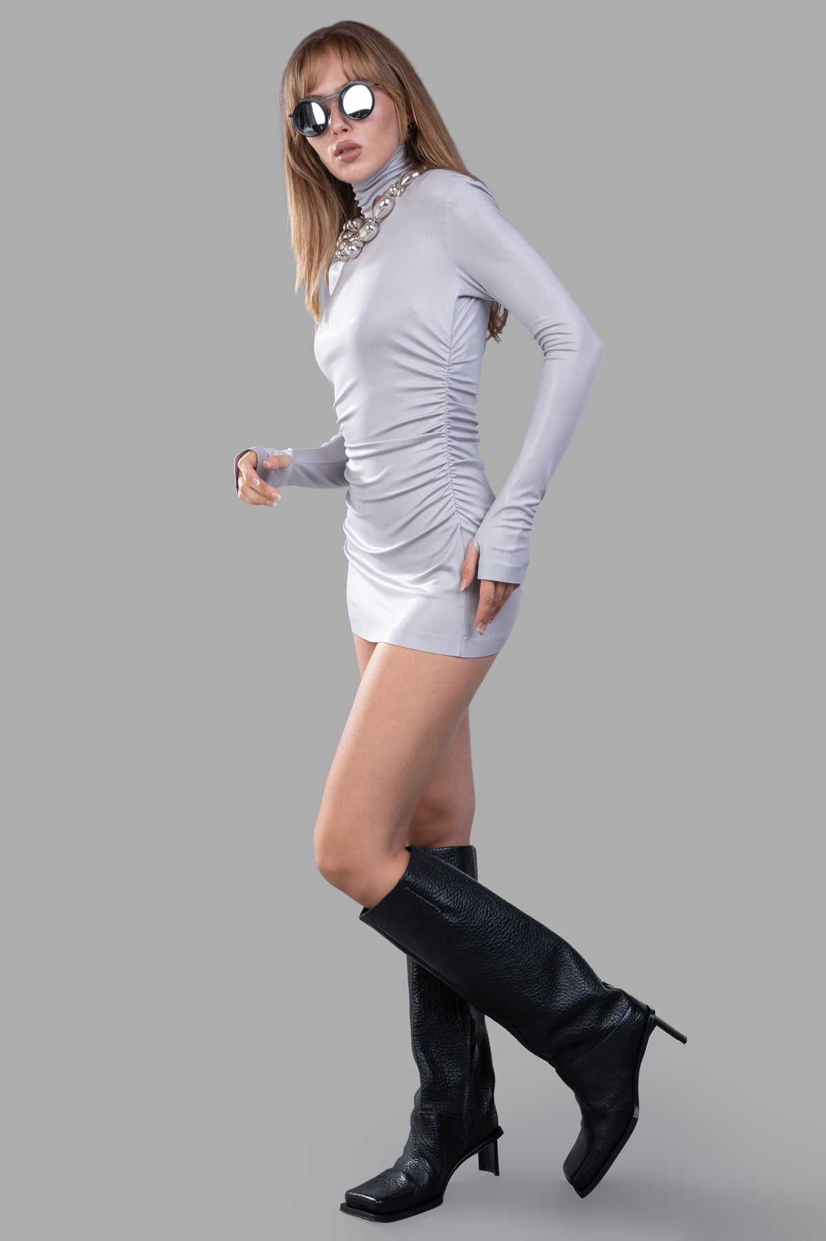 Norma Kamali Draped Mini Dress in silver, side view on model, long sleeves with thumb holes, black boots.