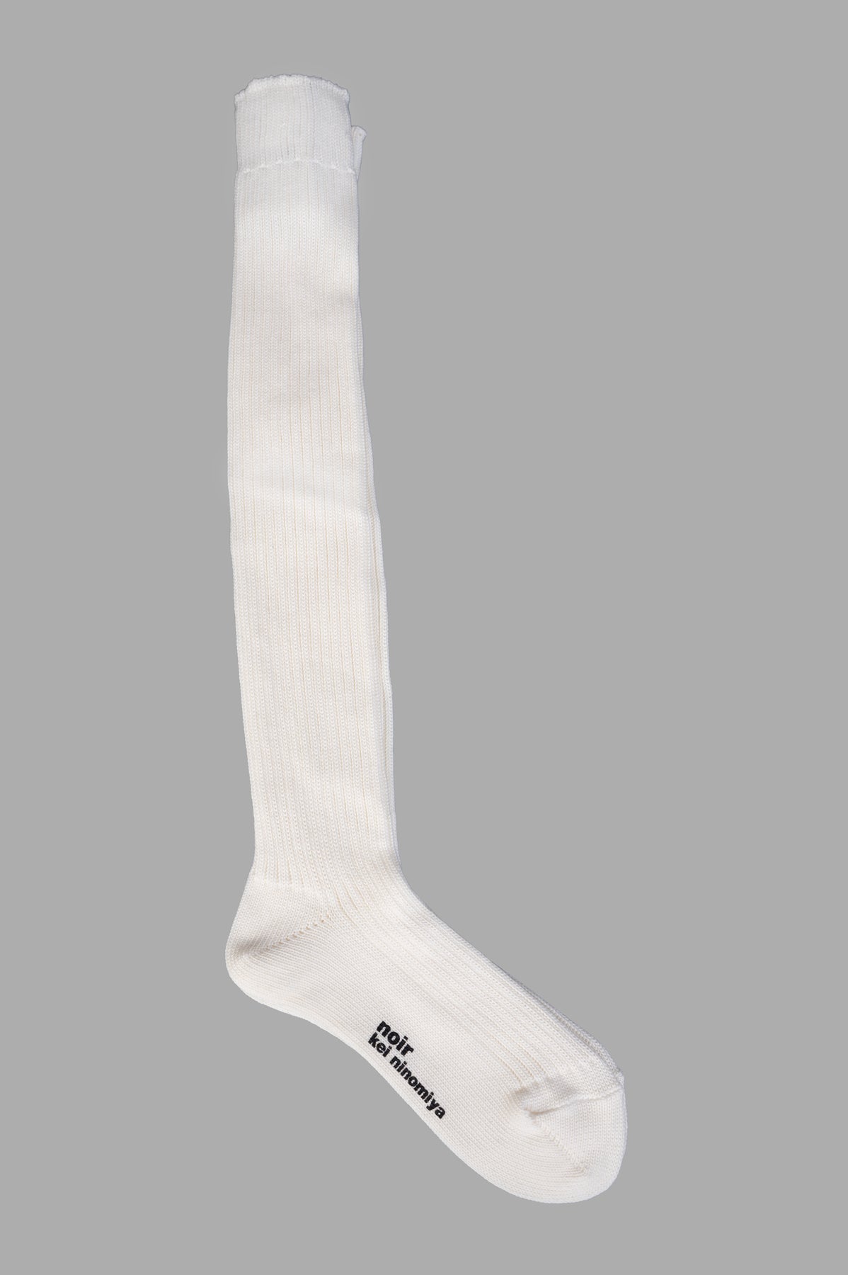 Noir Kei Ninomiya knee high socks, white ribbed texture, logo at sole, bold style for fashion enthusiasts.