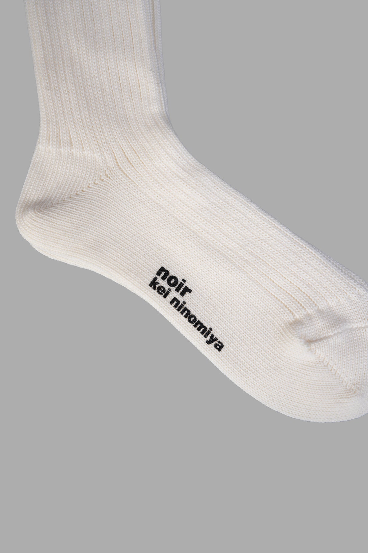 Noir Kei Ninomiya Knee High Socks in white feature ribbed texture and logo detail on the bottom for a bold fashion statement.