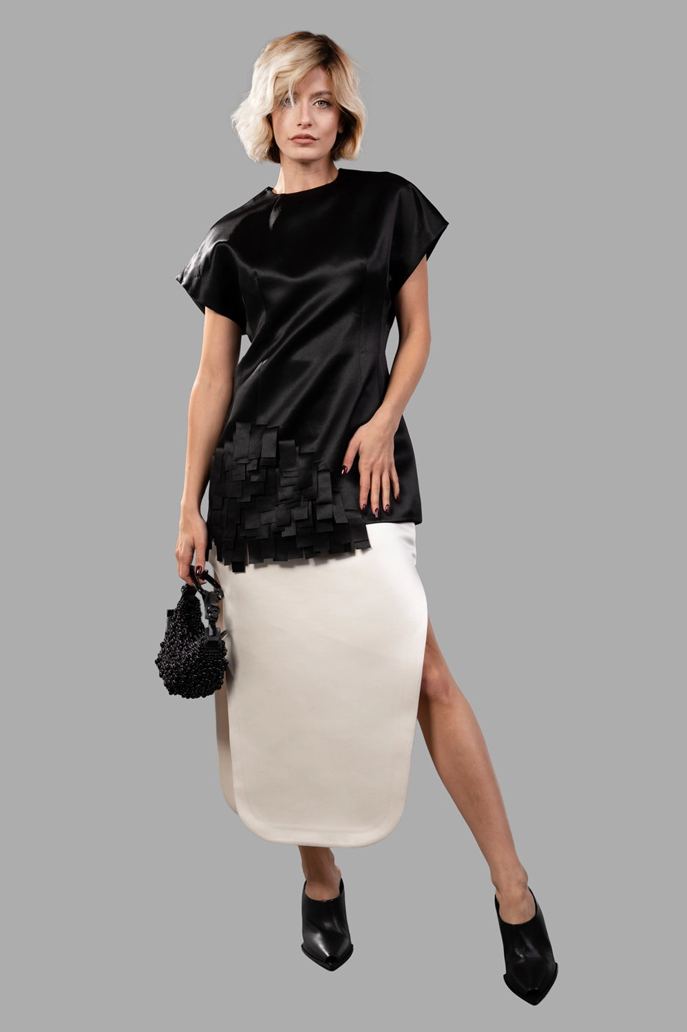 Full-length view of black satin top with wide neckline, cap sleeves and layered rectangle appliqué hem, styled with ivory skirt