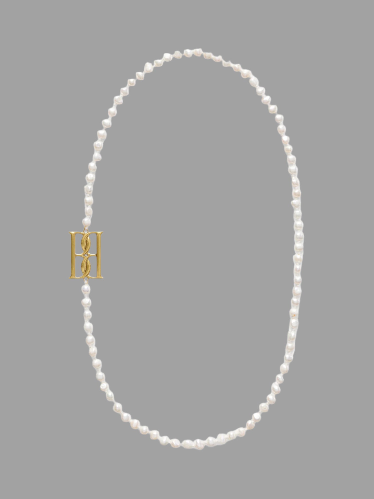 By Malene Birger Chakallo Monogram Necklace in white freshwater pearls with a gold monogram on a neutral background.