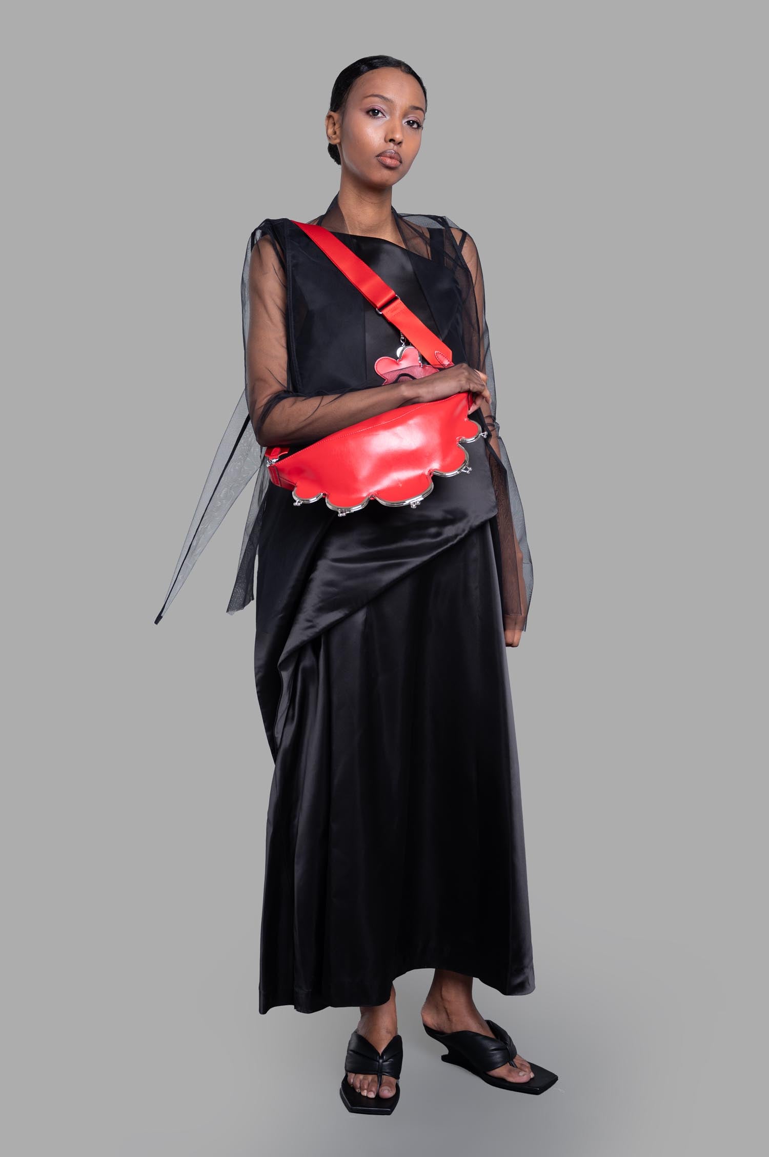Noir Kei Ninomiya Asymmetric Dress in Black, front view on model with sheer sleeves and a red bag accent.