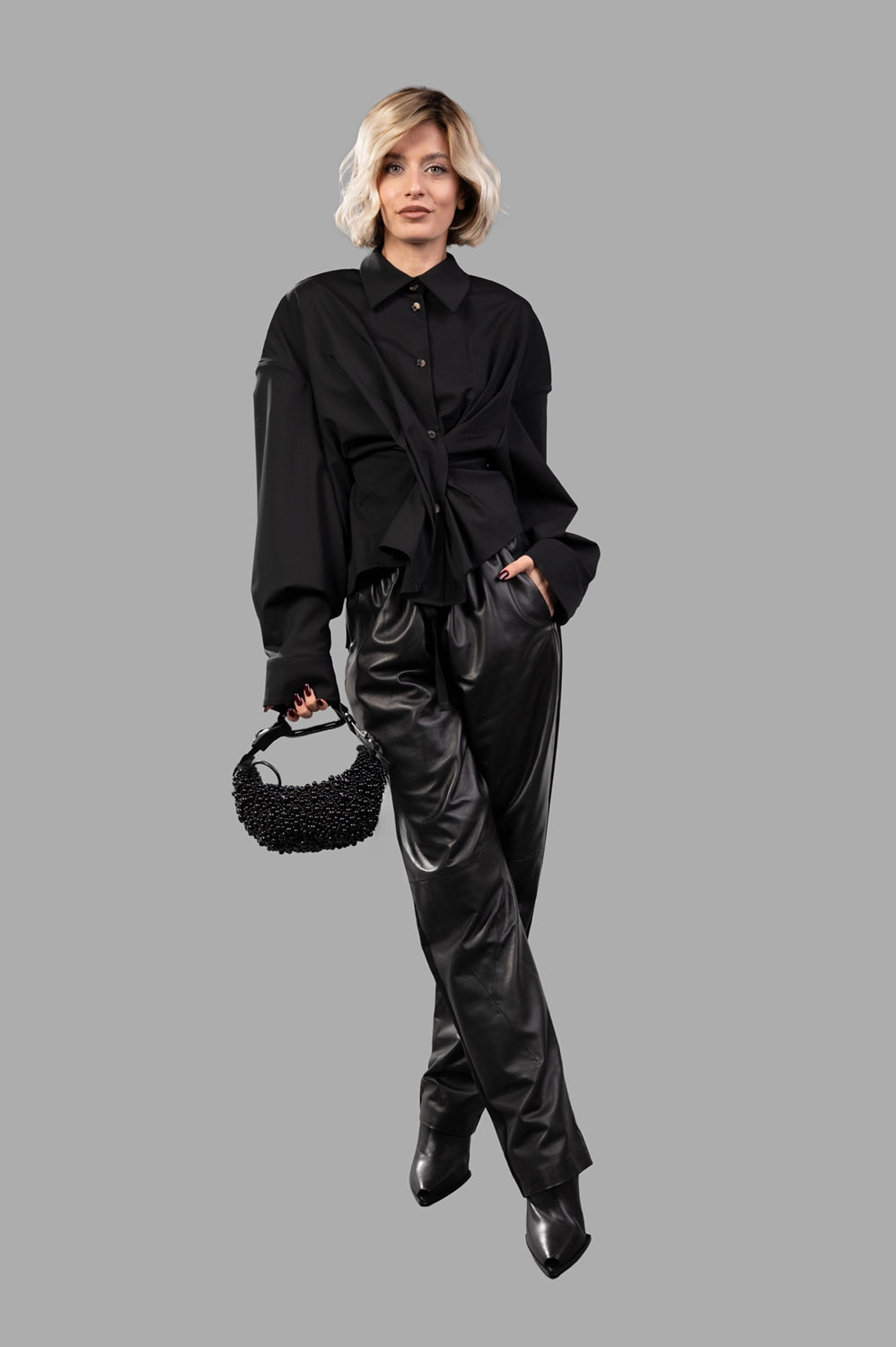 Full-length view of model wearing black twisted shirt with spread collar, dropped shoulders and draped waist, styled with black trousers