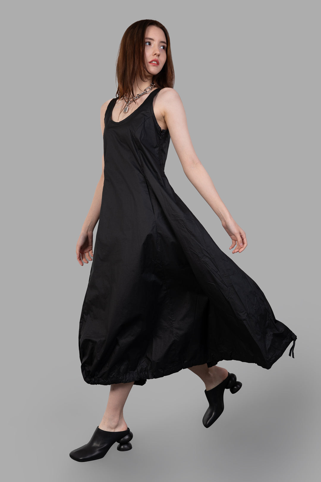 Masnada Balloon Dress; side view on-model, black sleeveless design with flowing hem, styled with unique black shoes.