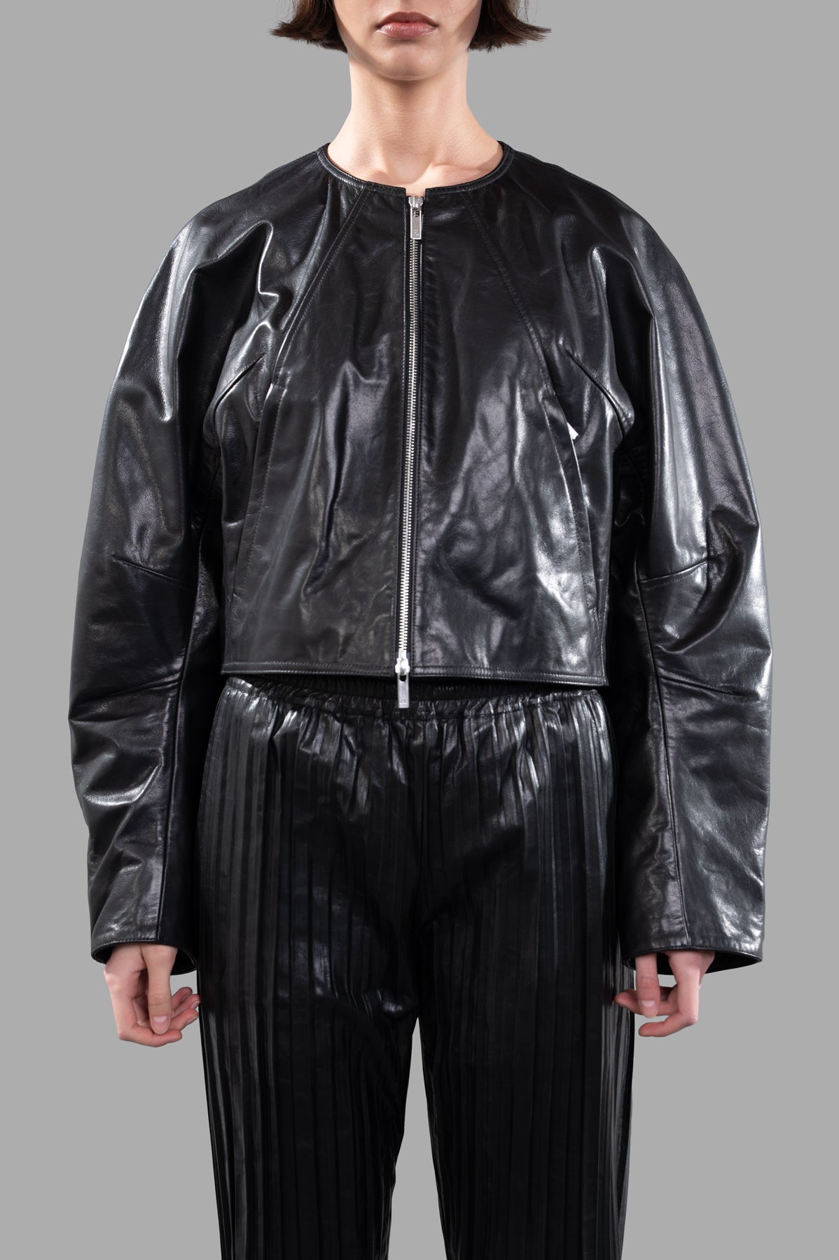 Helmut Lang Articulated Leather Jacket on model, featuring a cropped design and glossy finish, styled with pleated pants.