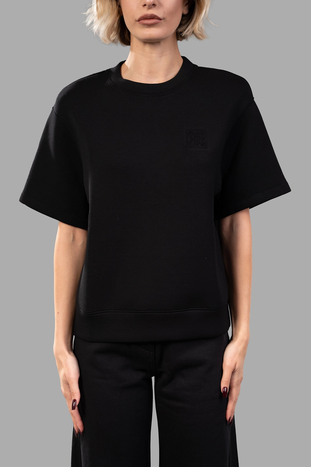 Front close-up of black short-sleeve sweatshirt showing crew neck, boxy fit, ribbed hem and tonal framed monogram at chest