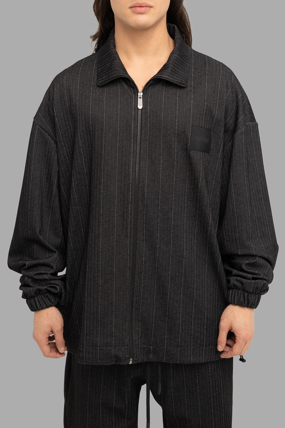 Gaëlle Paris men’s zip jacket in black, front view, pointed collar, chest logo patch and silver zip.